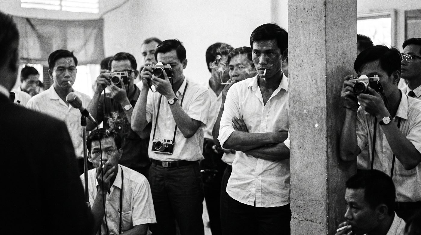 Hải standing at the edge of a press briefing, arms crossed, watching the room with a flat, unimpressed stare.