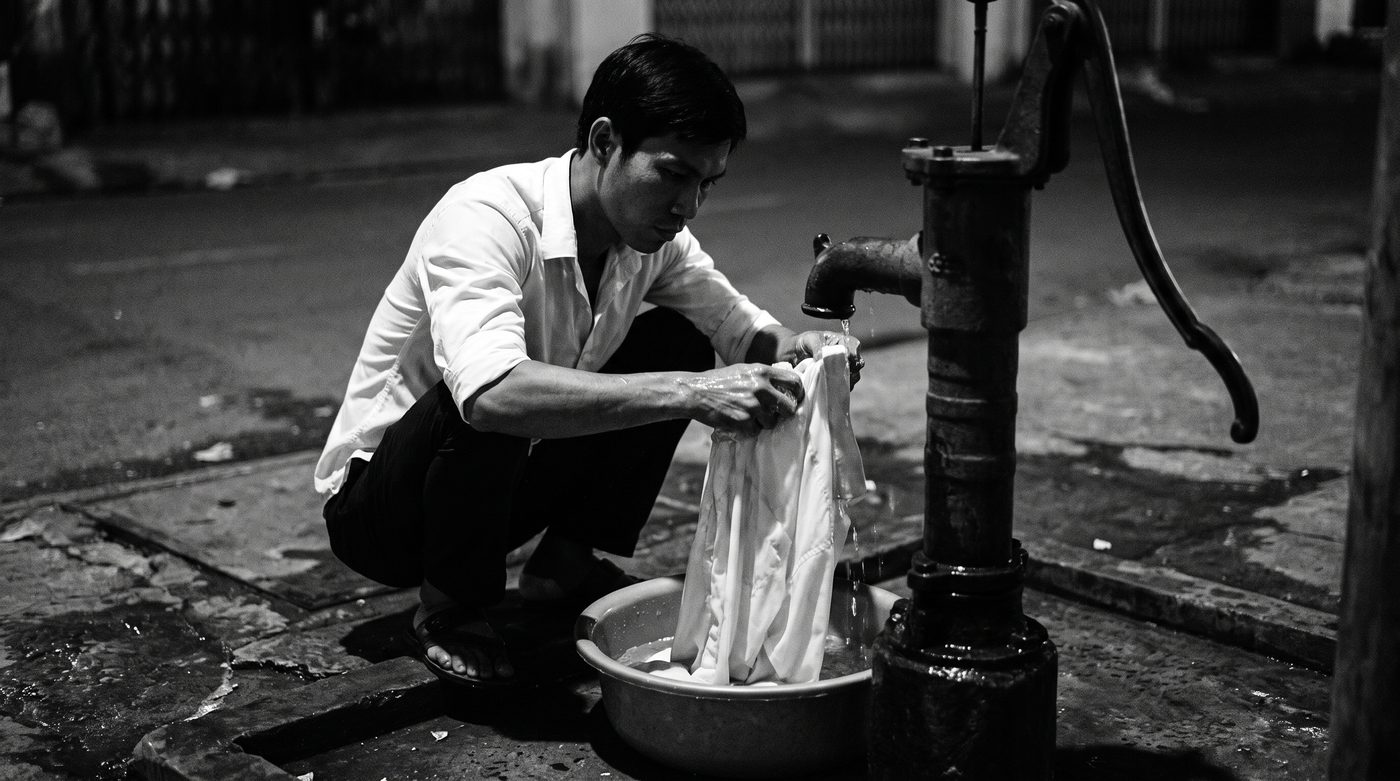 Hải washing a shirt at a pump, hands working the wet cloth, water running over his wrists.