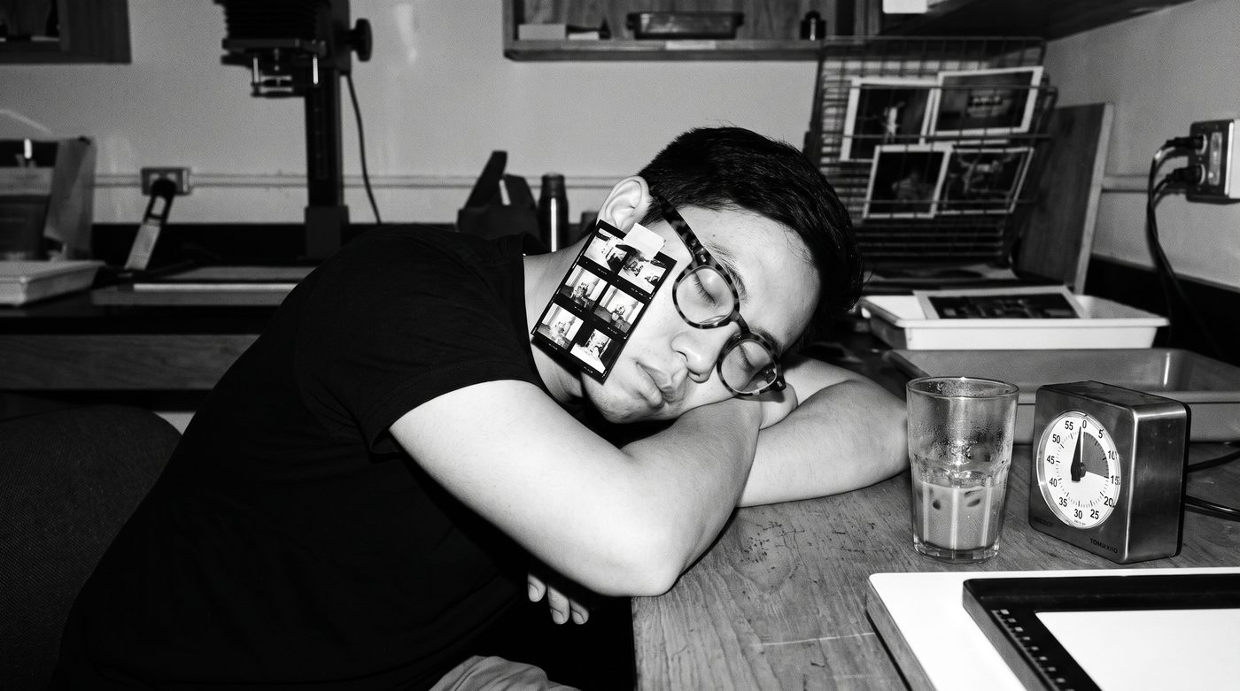 Minh Trần asleep at a desk, head down on his arms, a film timer glowing beside him.